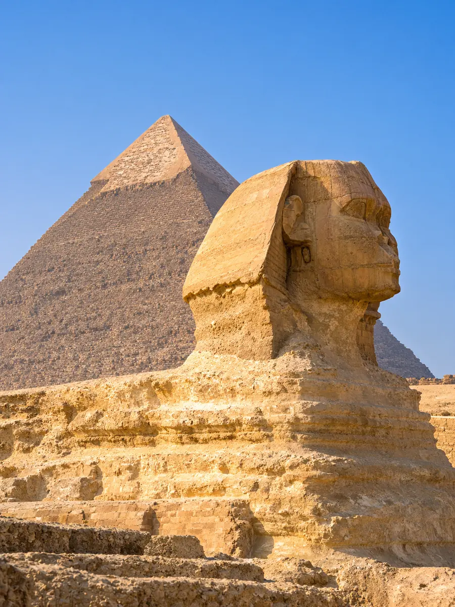 Die Große Sphinx von Gizeh von der Seite im warmen Morgenlicht, dahinter erhebt sich die Chephren-Pyramide unter blauem Himmel
